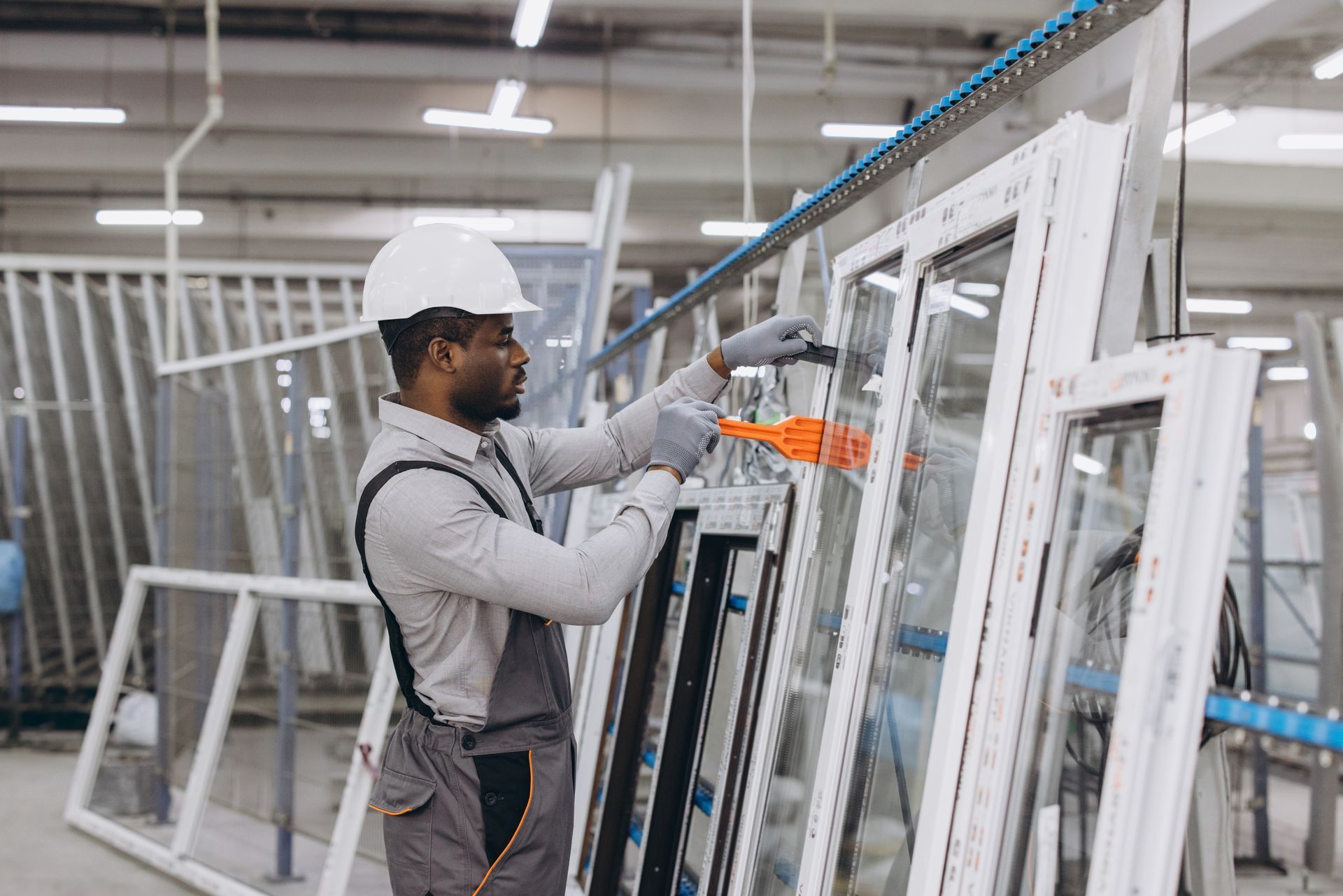 A worker in a white hard hat and gray uniform uses a tool to assemble a white vinyl window frame in a factory.