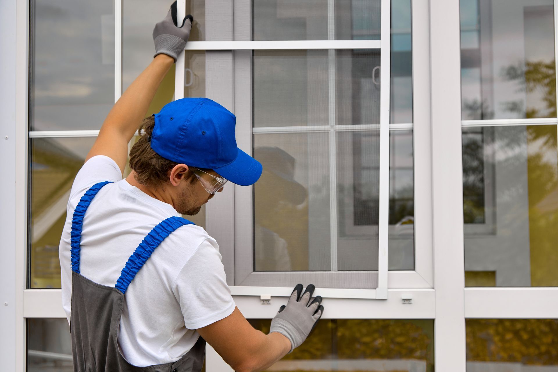 A worker in a blue cap, white shirt, and gray overalls installs a white window screen into a window frame.