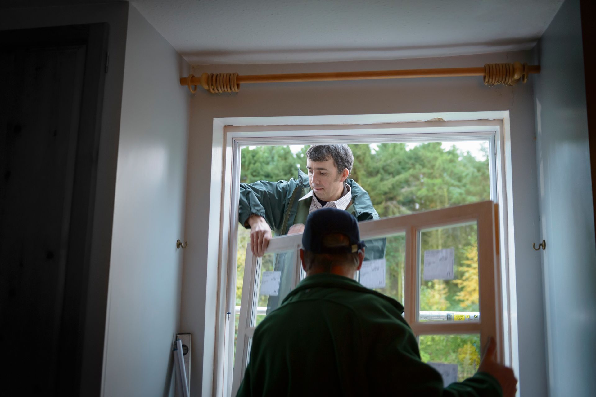 Two people working together to install a window frame in a home interior with trees visible outside.