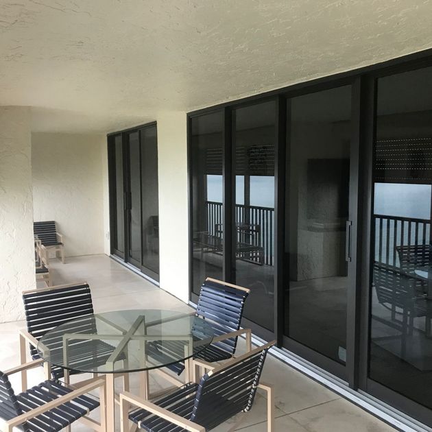 A glass-topped table with four modern, dark-slatted chairs on a balcony overlooking a railing and large sliding doors.