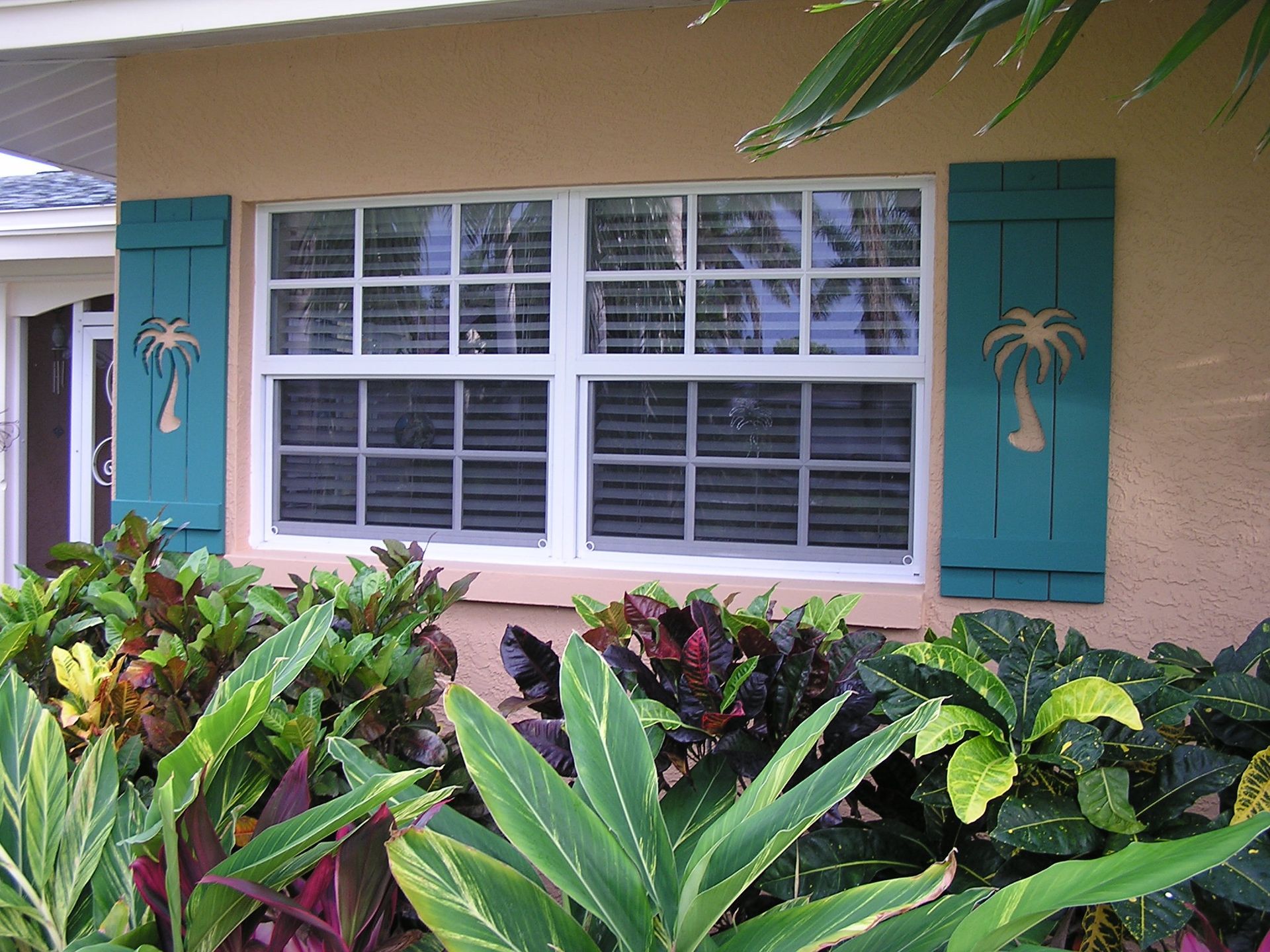 A white-framed window with teal shutters decorated with palm tree cutouts, surrounded by lush tropical plants.