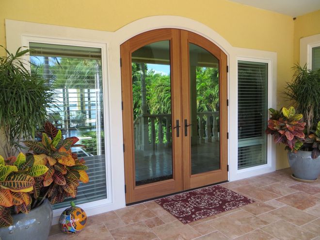 Double wooden doors with an arched top, flanked by side windows, on a porch with potted plants and tiled flooring.