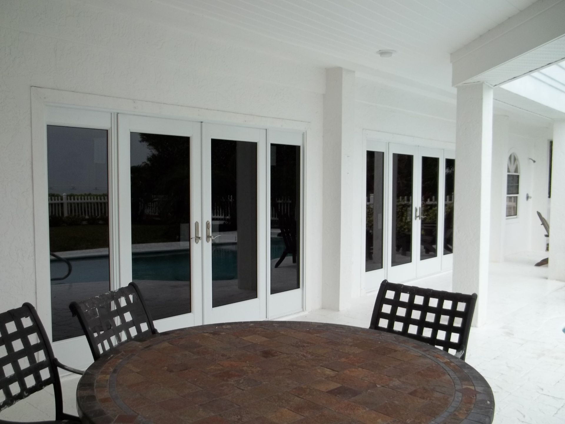 A patio with a stone-topped table and three chairs, overlooking a swimming pool through large white-framed glass doors.