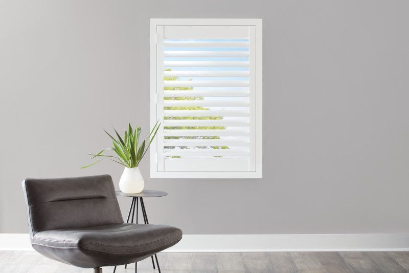 A charcoal armchair and a small side table with a potted plant sit before a window with white plantation shutters.