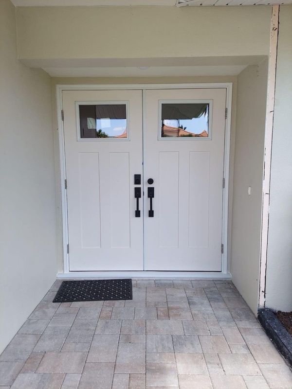 A white double entry door with rectangular windows and black handles, set against a light wall above a paved porch.