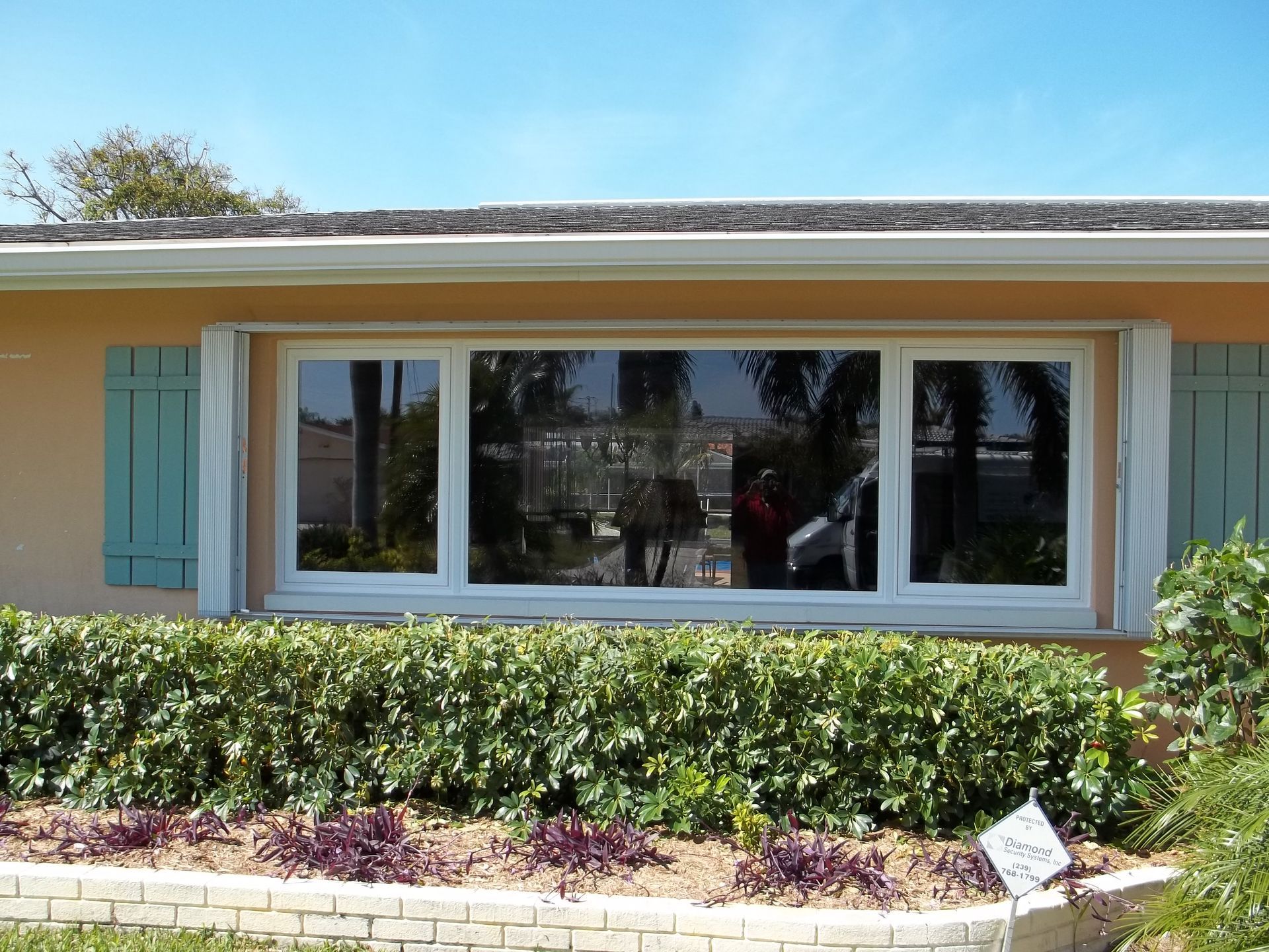 A tan house exterior with a large, three-paneled window, light blue shutters, and a manicured hedge in front.