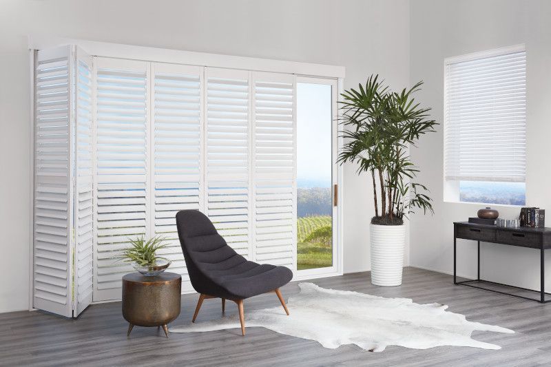 A modern room with white plantation shutters, a dark lounge chair, a potted palm, and a black console table.
