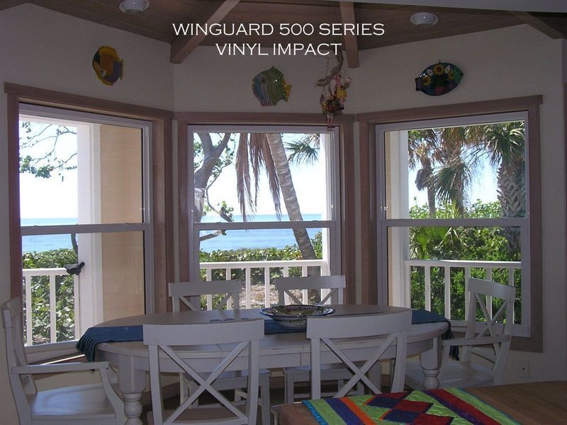 A dining room with white chairs, a table, and three large windows overlooking a beach, palm trees, and the ocean.