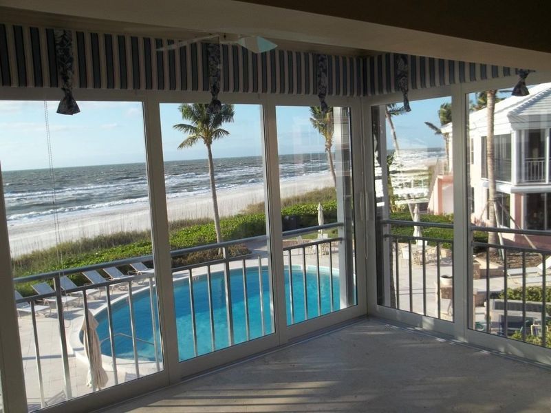A balcony with a striped valance overlooking a swimming pool, sandy beach, and ocean under a bright sky.