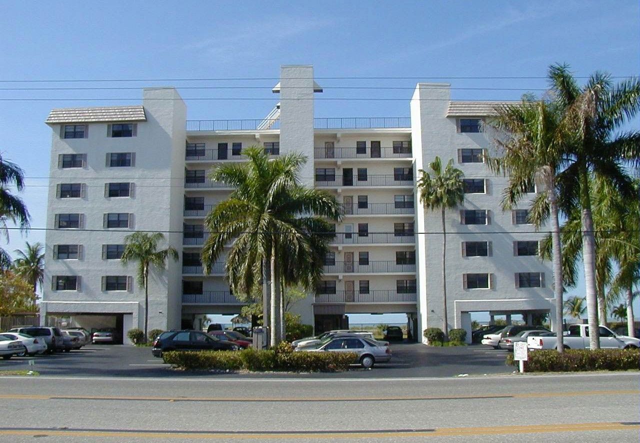 A multi-story, white apartment building with palm trees and parked cars in front under a clear blue sky.