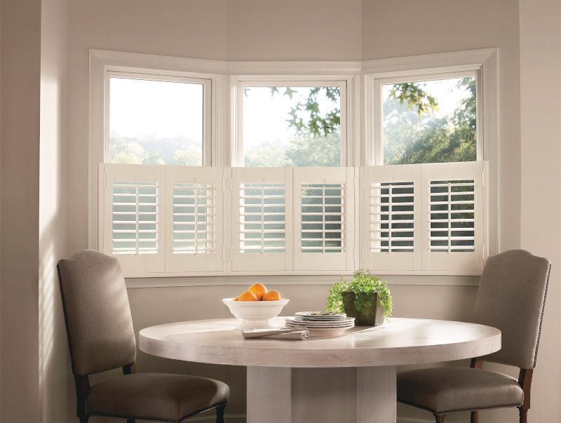 A dining nook with a bay window, white café shutters, a round white table, and two gray upholstered chairs.
