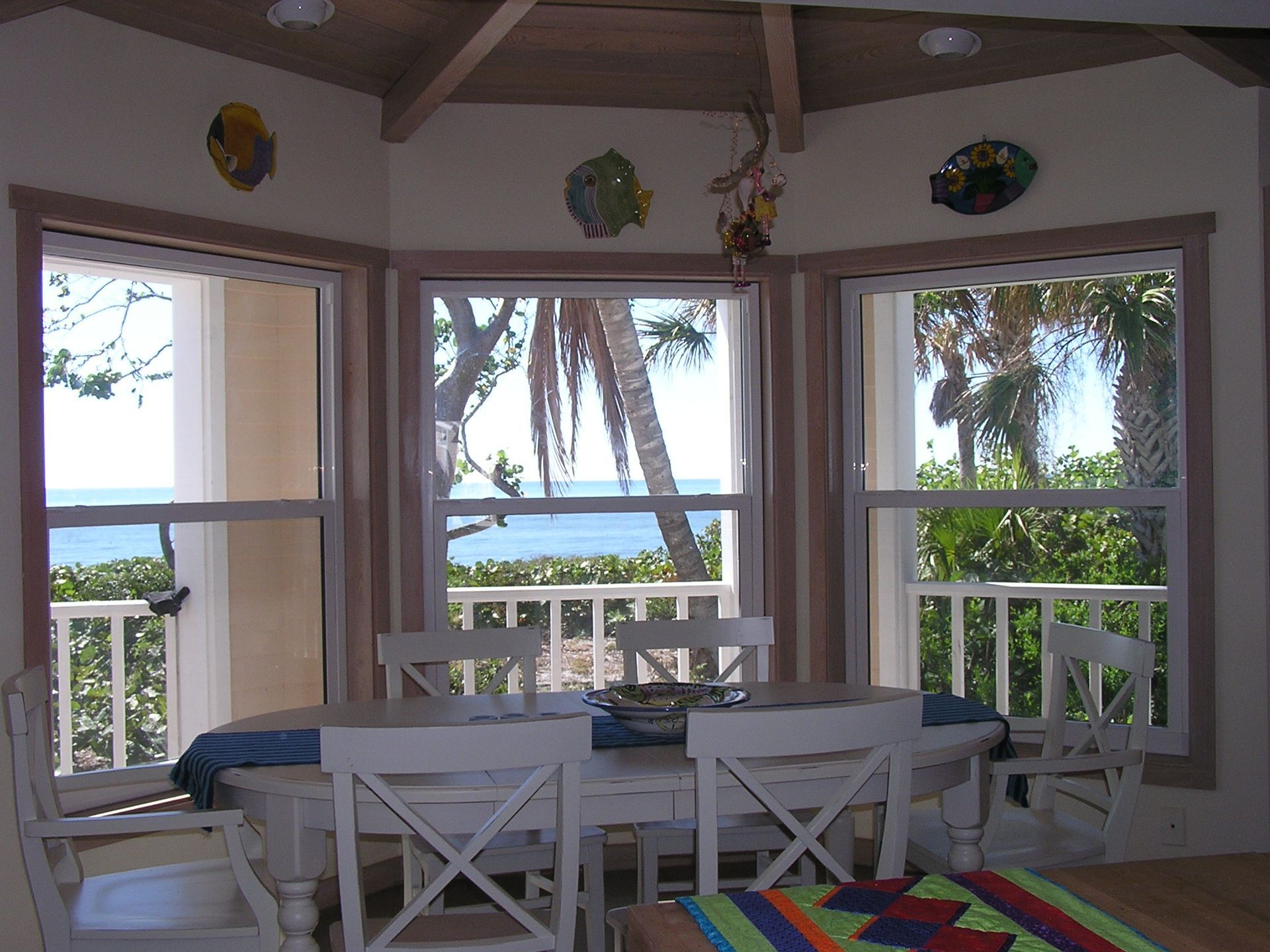 A white dining table with four chairs sits in a sunlit room featuring bay windows overlooking the ocean and palm trees.