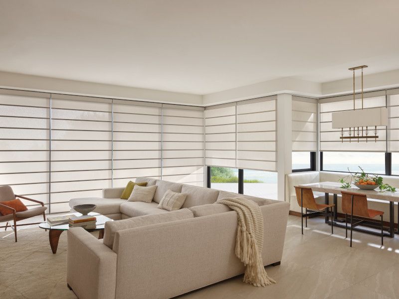 A modern living room with a large beige sectional sofa, a coffee table, and a dining area with window shades.