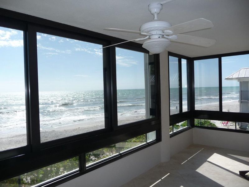 A sunroom with large windows overlooking a sandy beach and ocean, featuring a white ceiling fan.
