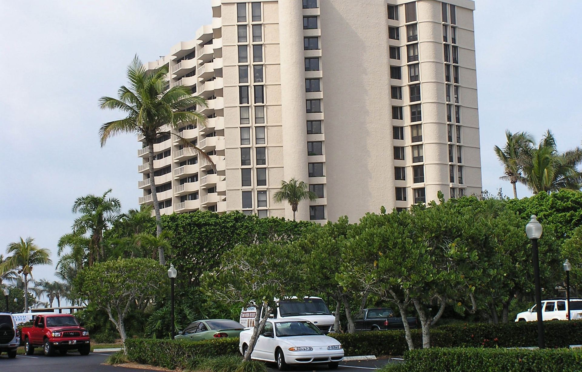 A tall, beige multi-story apartment building rises behind lush green trees and a parking lot with several parked cars.