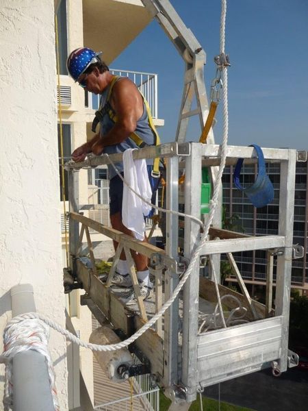A worker in a safety harness on a suspended platform performs exterior maintenance on a high-rise building.