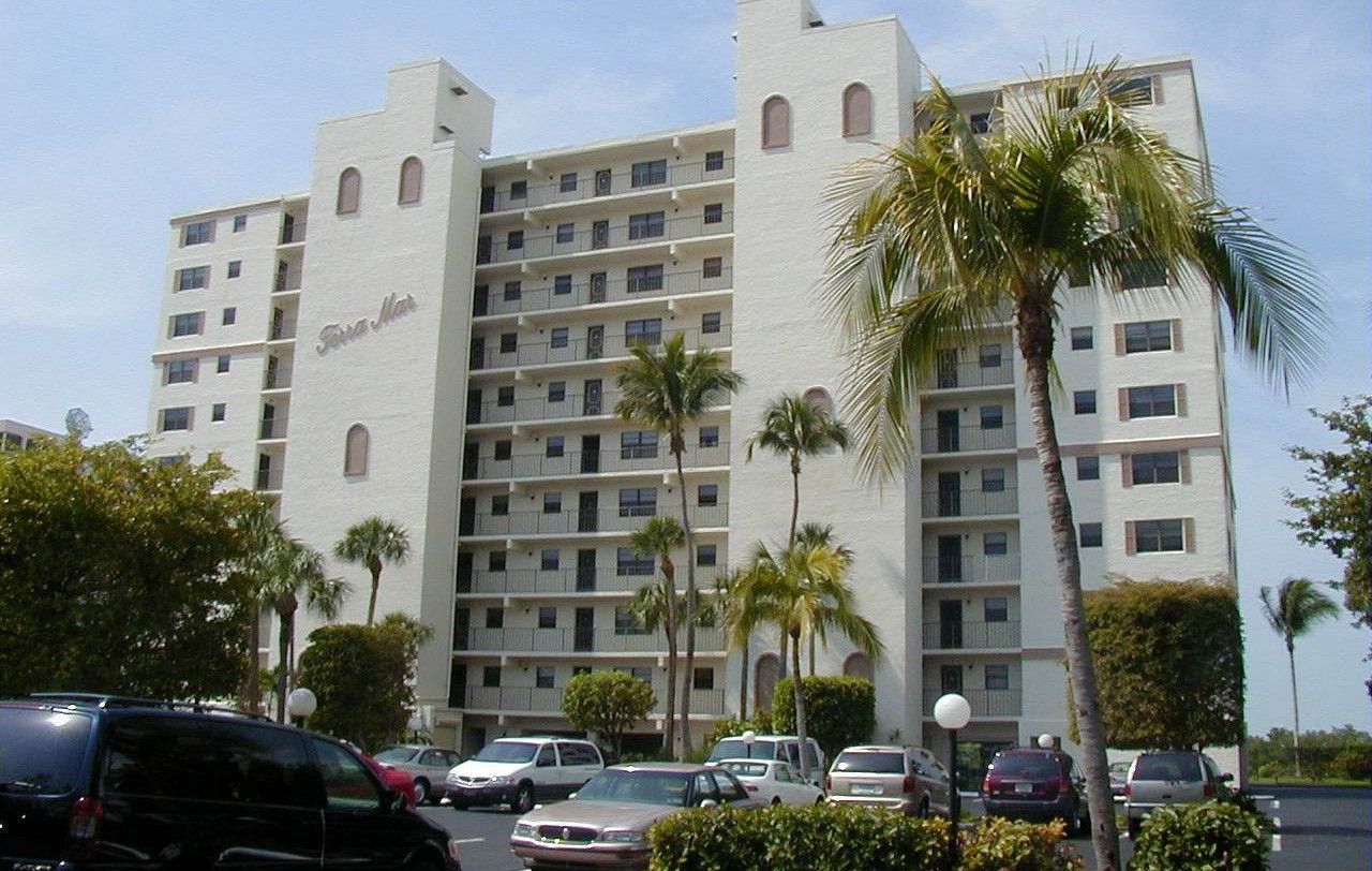 A tall, white apartment building with balconies and arched windows, surrounded by palm trees and a parking lot.