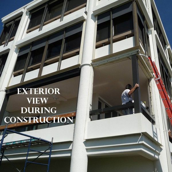 A person works on the balcony of a multi-story white building during construction, with a ladder leaned against the side.