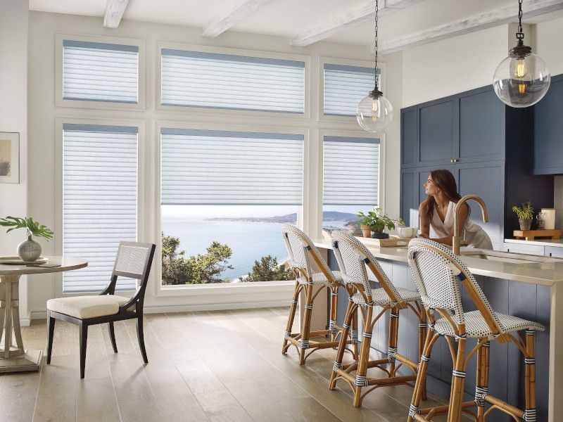 A sunlit kitchen featuring blue cabinetry, rattan stools at an island, and large windows with blinds overlooking the ocean.