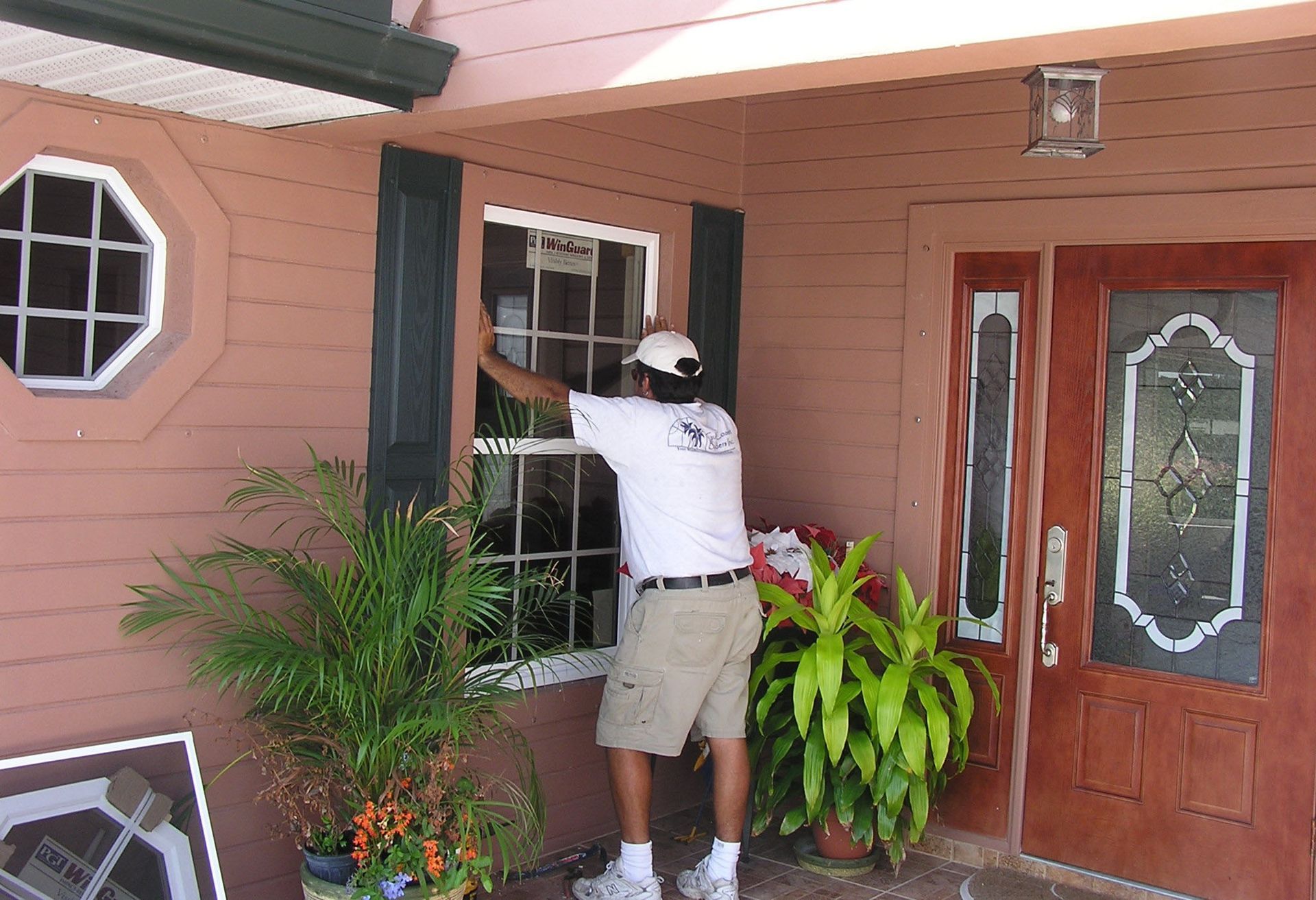 A person in a white shirt and khaki shorts stands outside a home, installing or repairing a window near the front door.