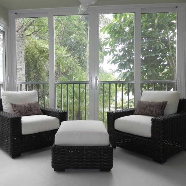 Two dark wicker armchairs with white cushions and brown pillows face a matching white ottoman in front of a glass wall.