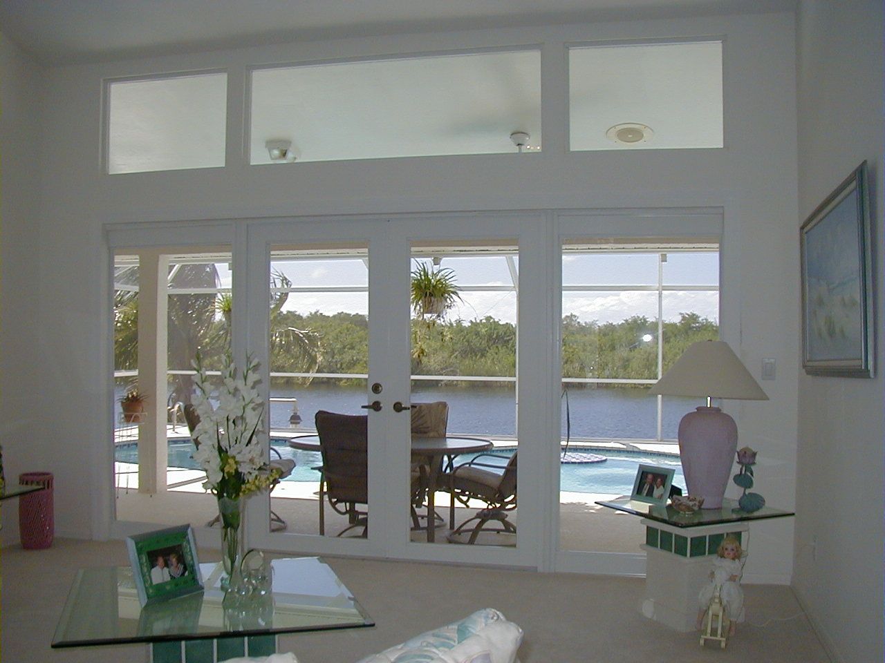A bright living room looks out through a wide glass door onto a backyard pool and river with trees in the distance.