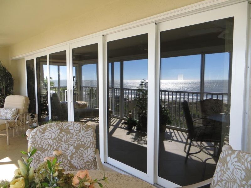 White-framed sliding glass doors open onto a balcony with a view of the ocean, seen from an indoor dining area.