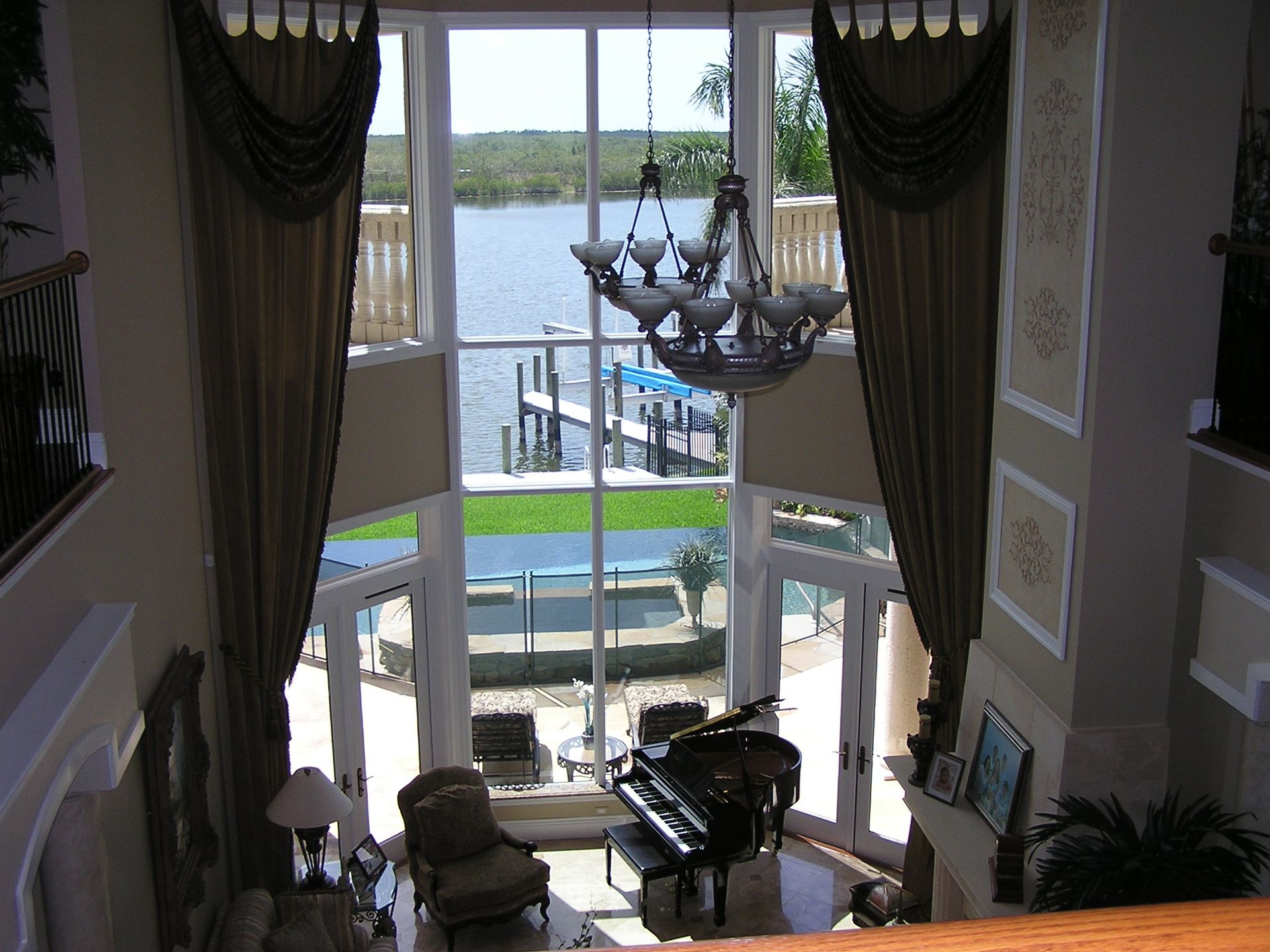 A grand room with floor-to-ceiling windows looking out over a dock and water, featuring a piano and chandelier.