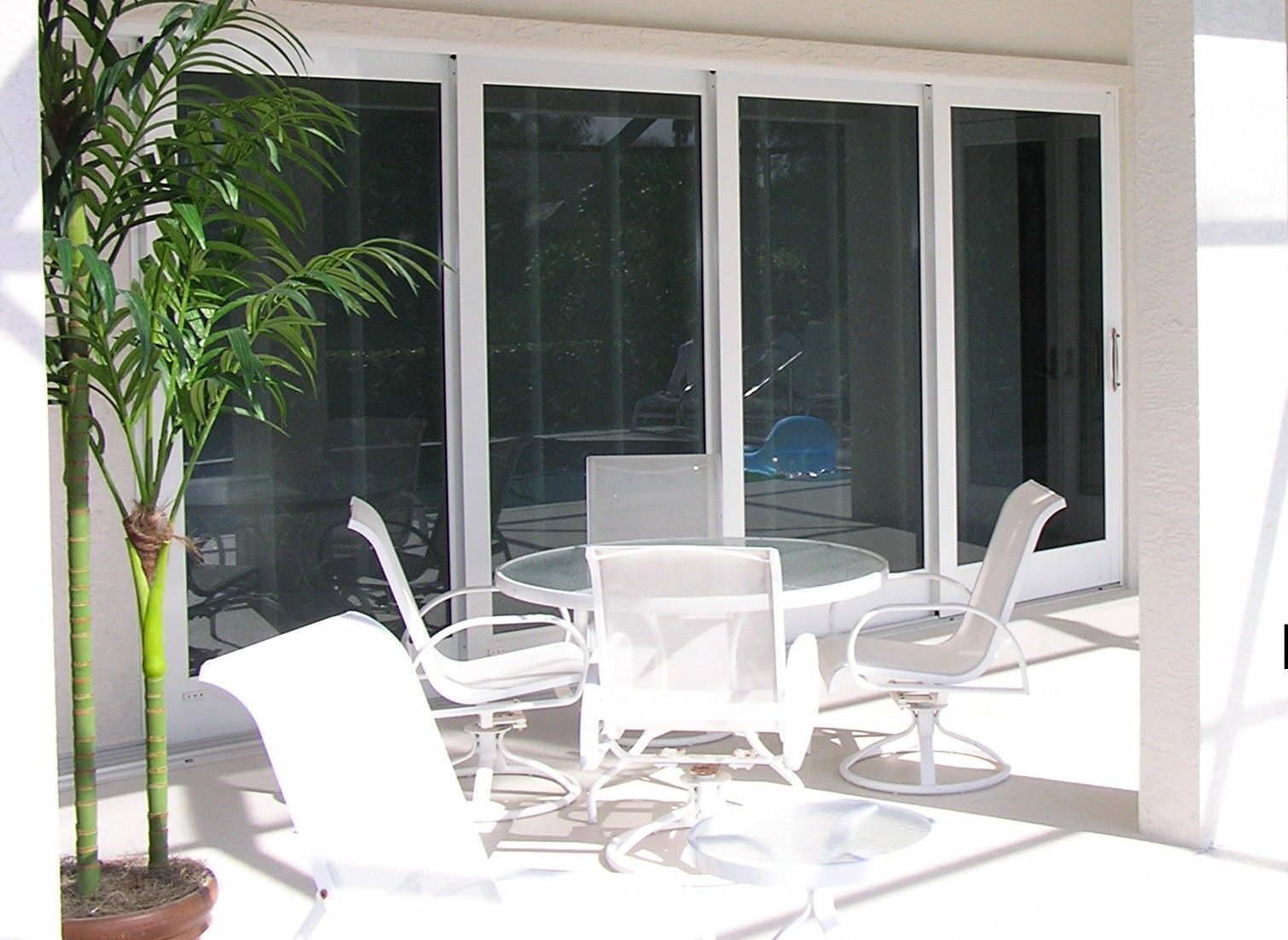A patio with a glass-topped table and chairs, situated in front of a multi-panel sliding glass door, next to a potted palm.