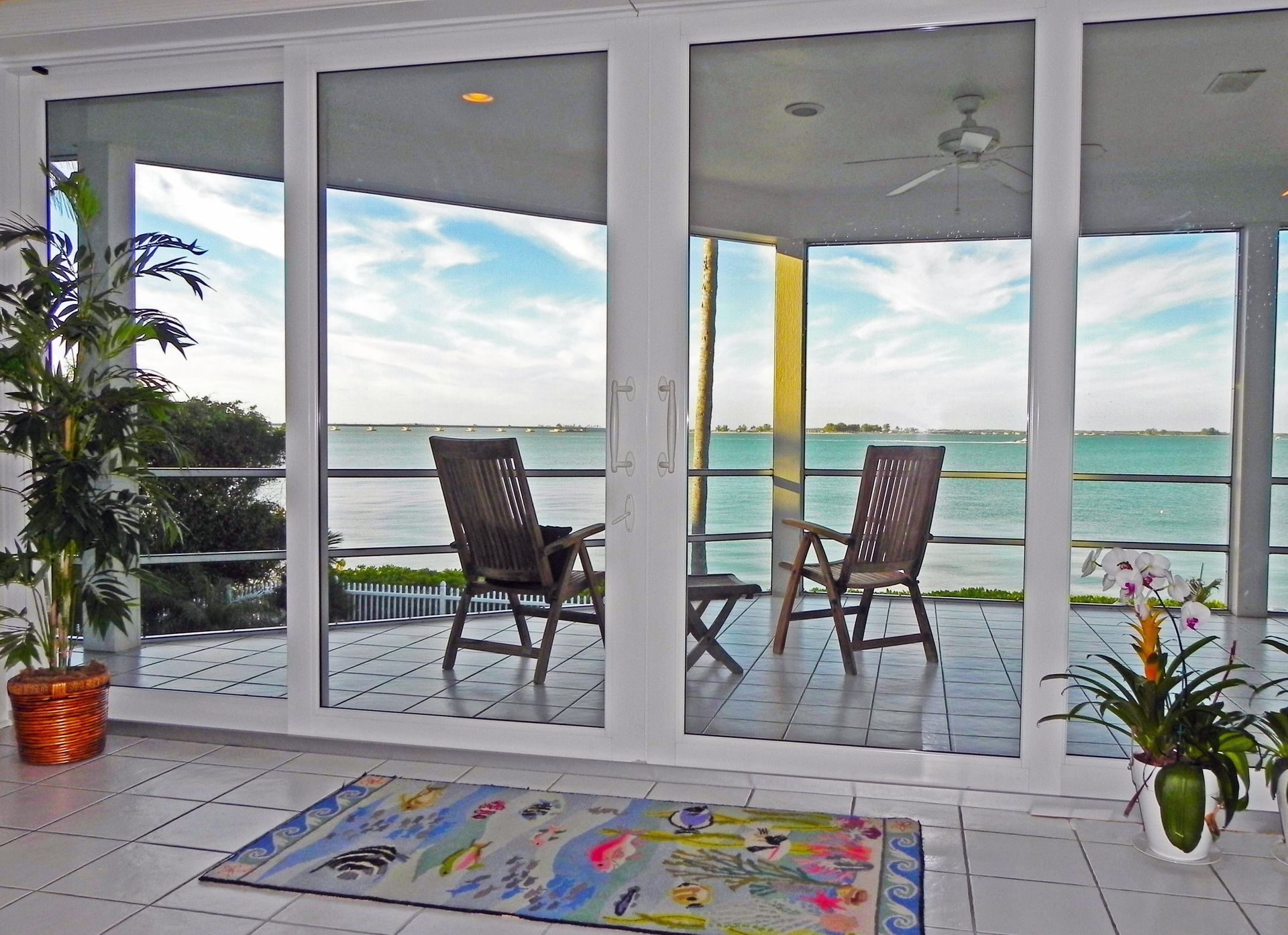 A sunlit room with tiled floors looking out through glass doors to two chairs on a patio overlooking a calm ocean.