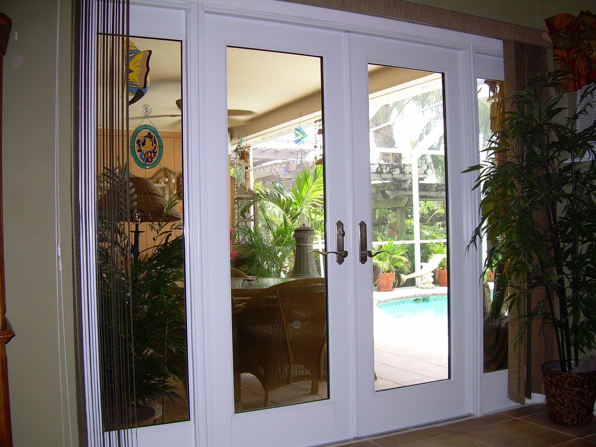 White double glass doors looking out onto a patio and swimming pool with lush green plants inside and outside.