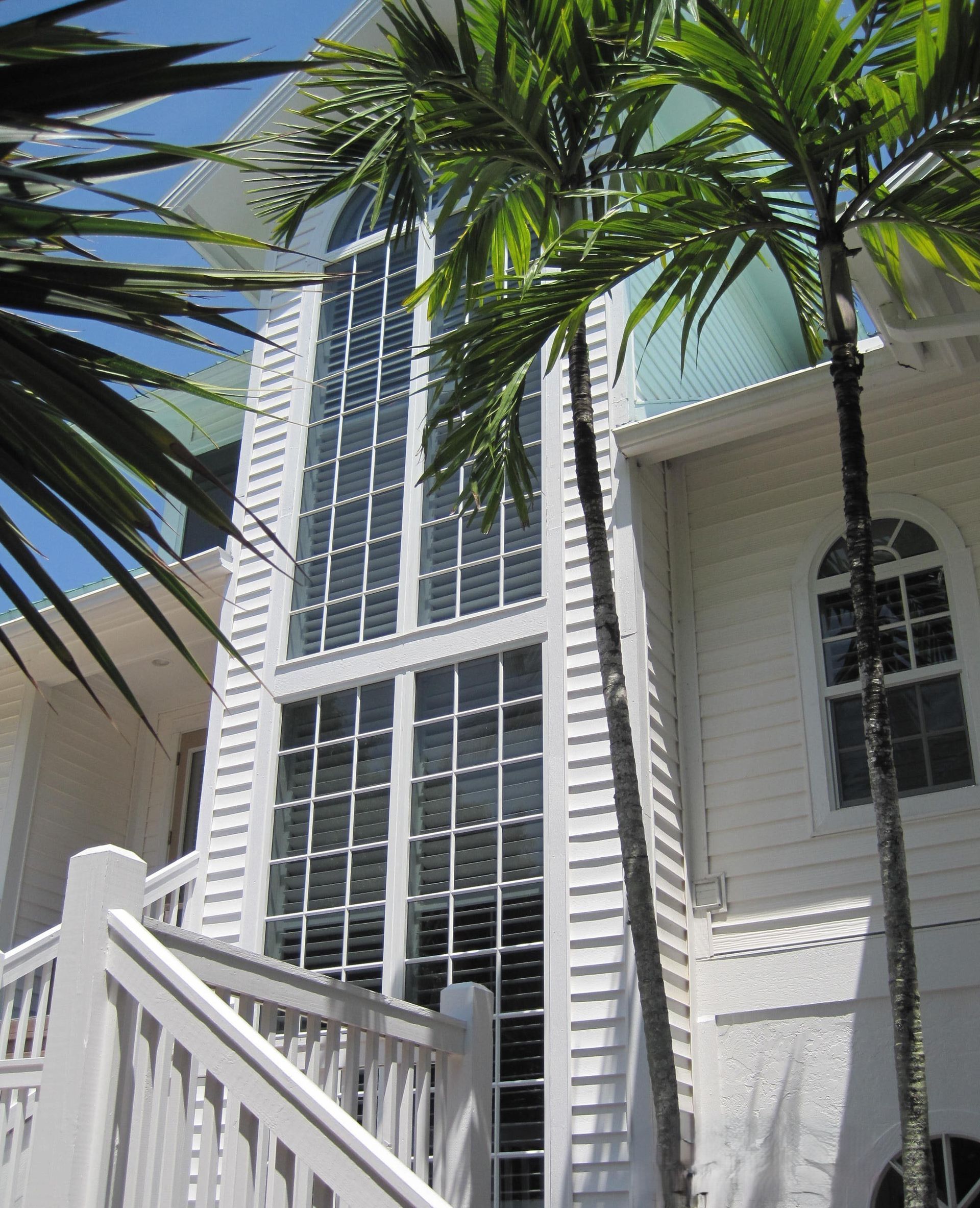 A white house with a tall vertical window bank, white stairs, and palm trees against a bright blue sky.