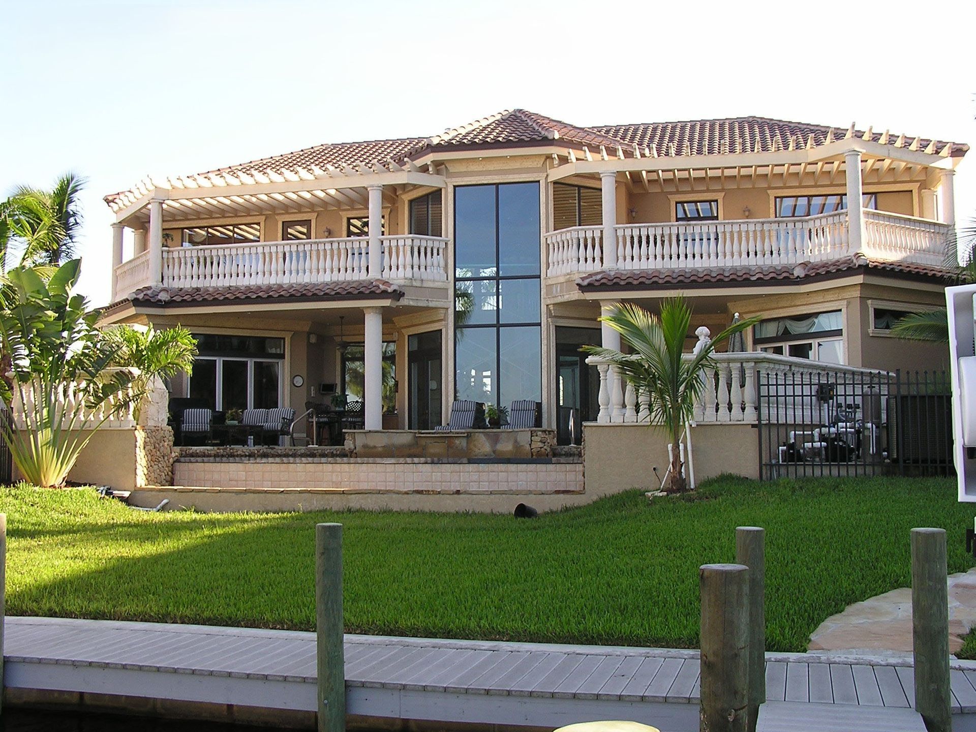A large, two-story beige waterfront house with a balcony, tiled roof, and a central vertical window panel.
