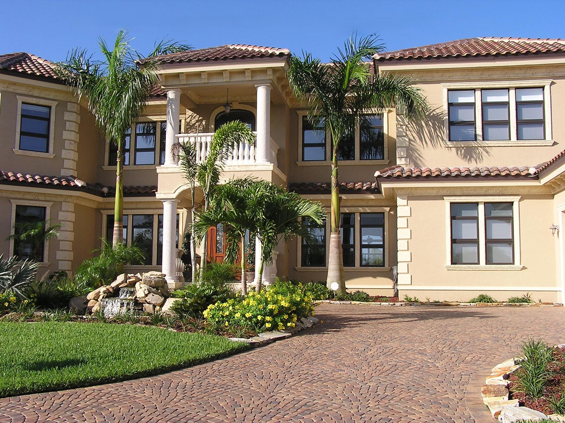 A large, two-story, tan stucco luxury home with a tile roof, columns at the entrance, and a paved driveway.