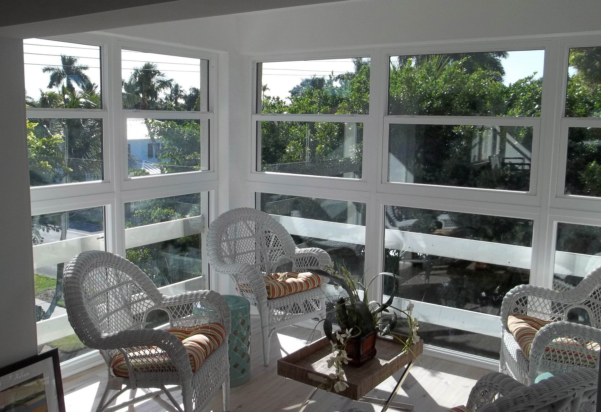 Sunroom with white wicker chairs, a small table, and large windows overlooking a lush, sunny yard.