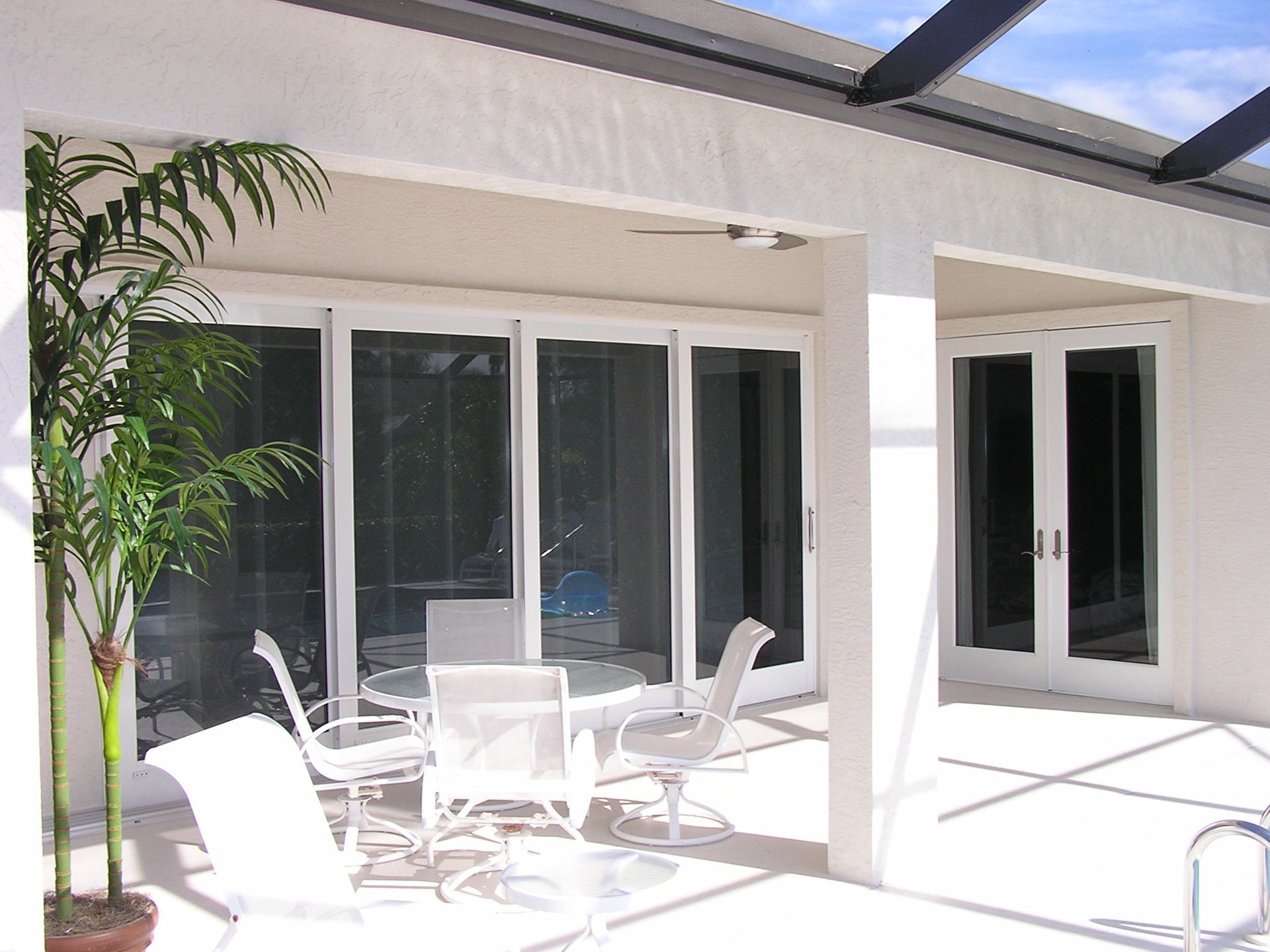 A white patio with a glass-topped table and chairs, situated under a covered walkway next to sliding glass doors.