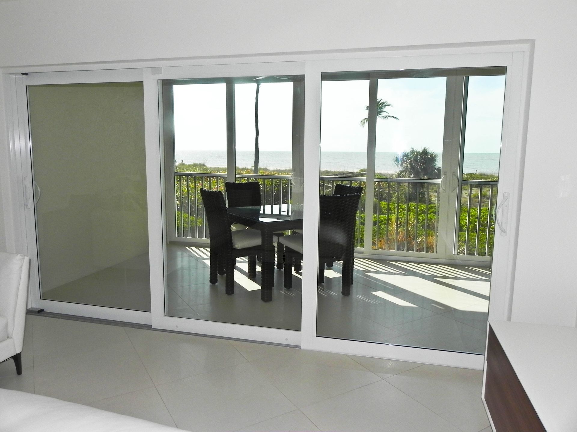A set of white sliding glass doors opening to a balcony with dark dining furniture and a view of the ocean.