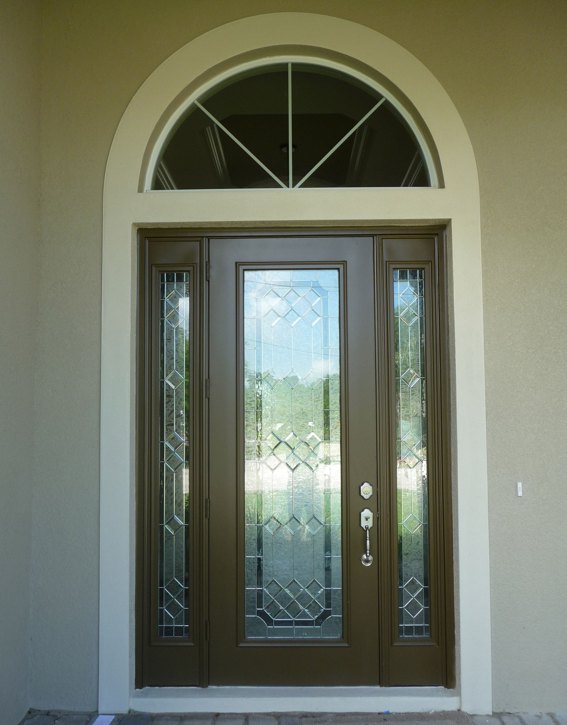 A brown front door with glass panels and sidelights, topped by a decorative arched transom window, set in a tan wall.