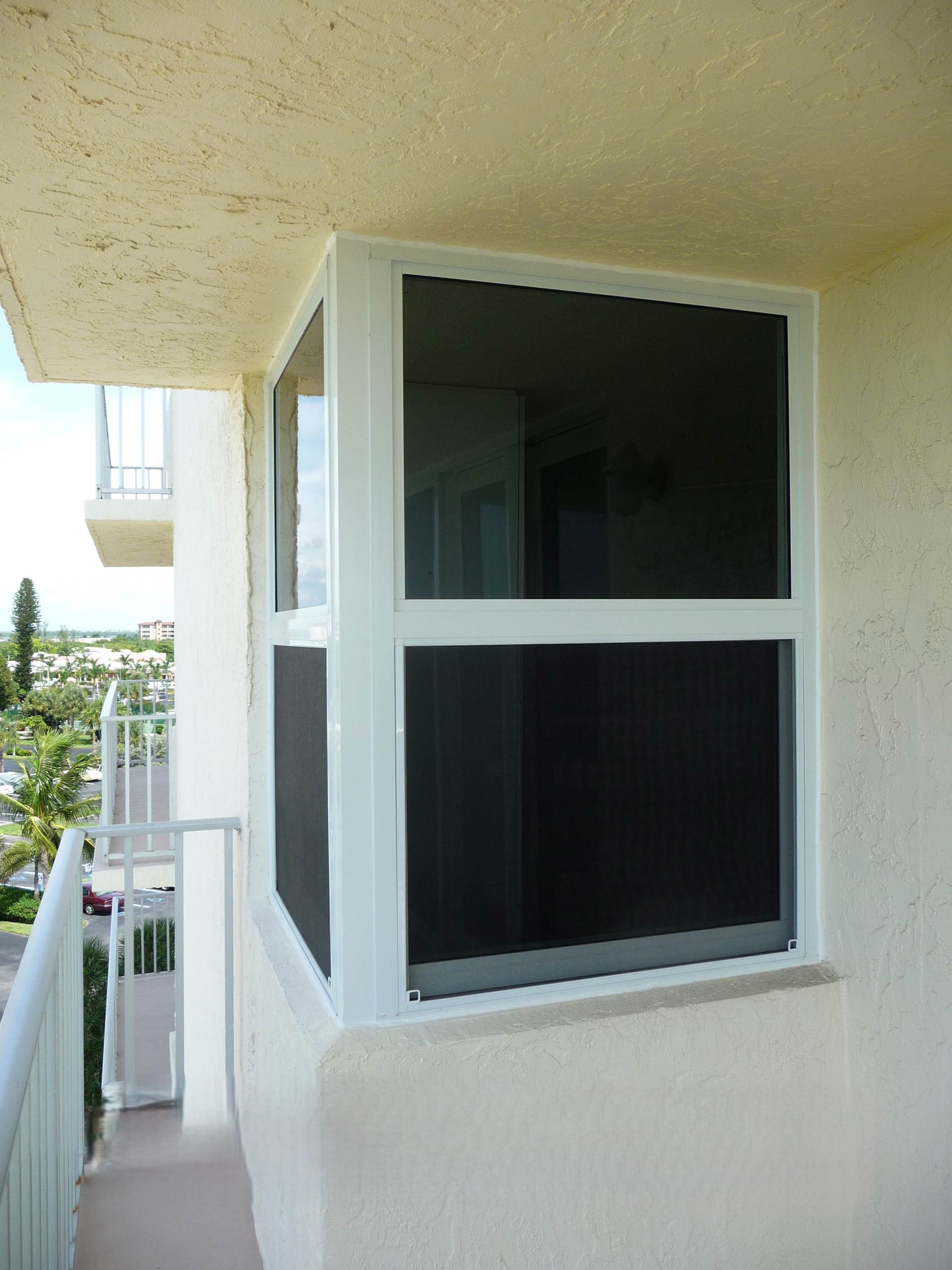 White window frame with black mesh screens installed on an exterior balcony wall of a stucco building.