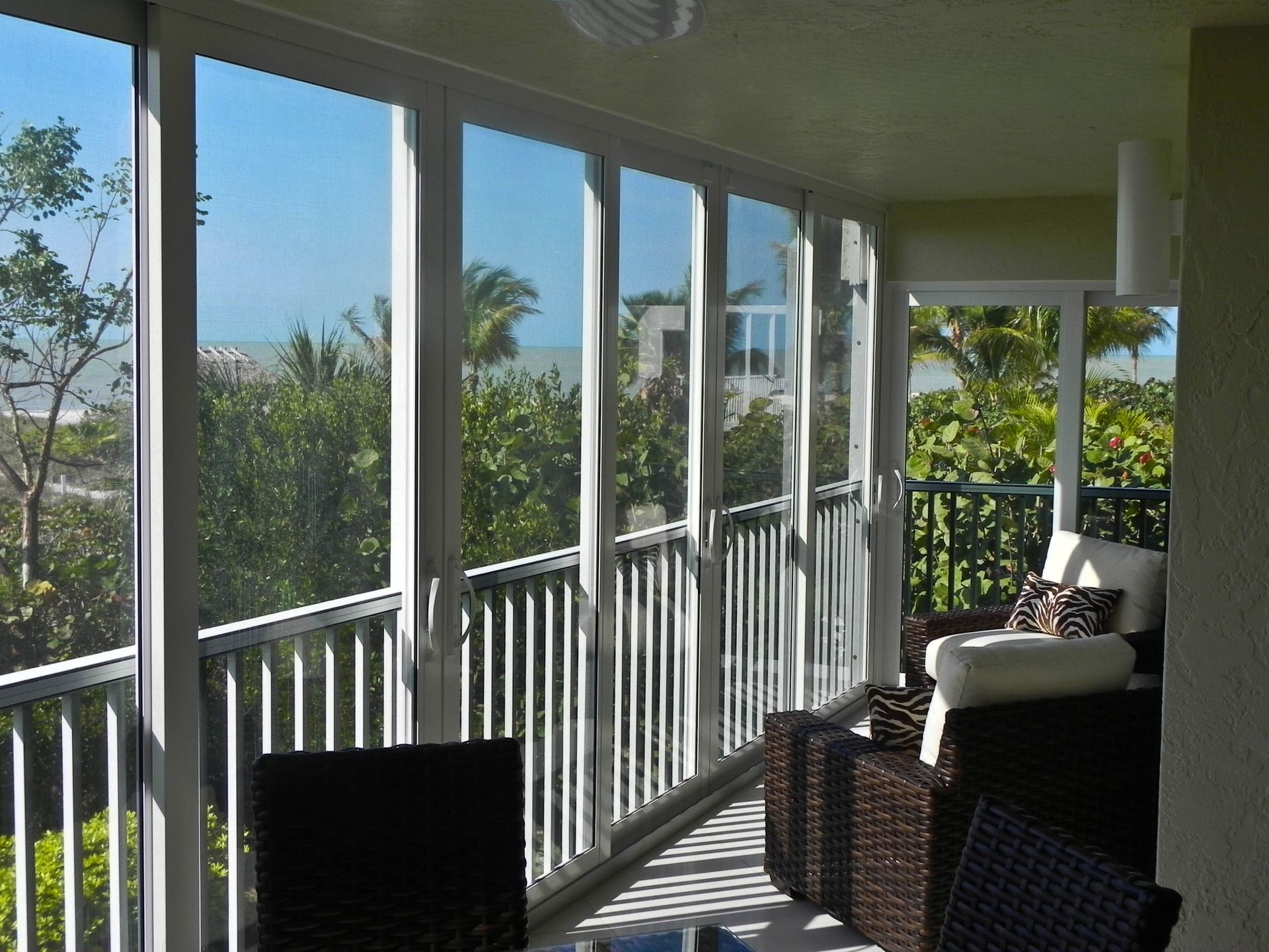 A sunroom balcony with large glass windows, white railings, and wicker furniture overlooking a lush, tropical beach view.