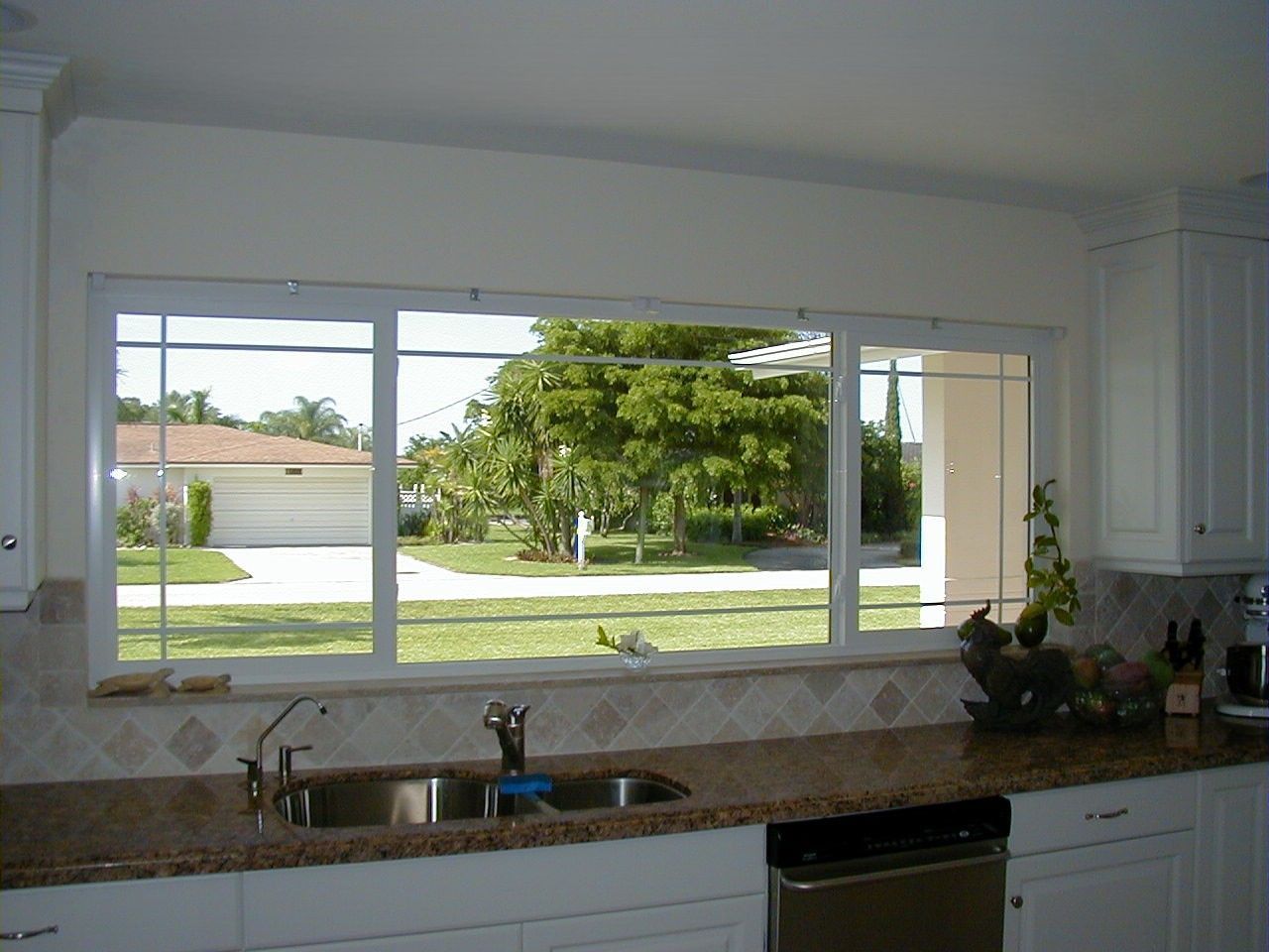 A wide kitchen window overlooking a residential front yard with a driveway, lawn, and mature trees.
