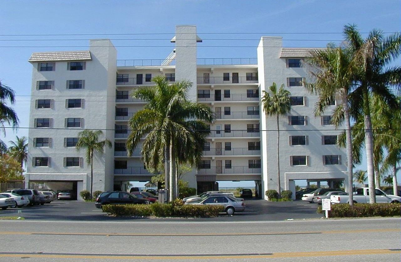 A multi-story white residential building with balconies, palm trees, and a parking level, set under a blue sky.