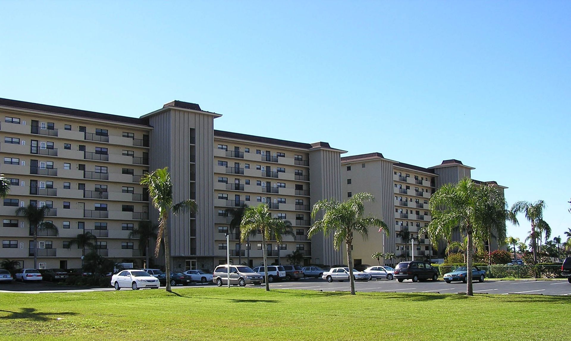 A multi-story apartment building with beige walls and balconies sits behind a lawn and parking lot under a blue sky.