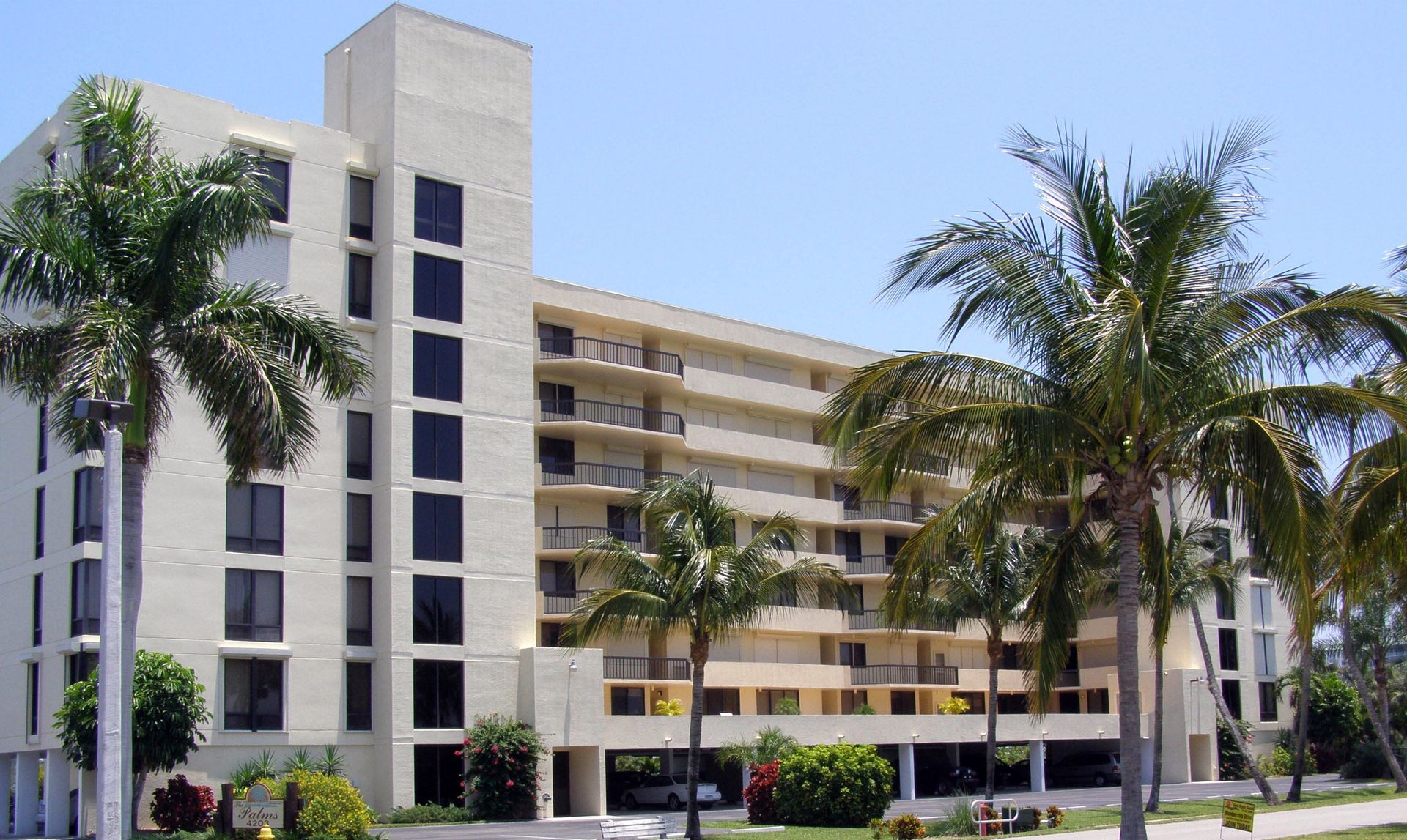 A light-colored multi-story apartment building with balconies and dark windows, surrounded by palm trees under a blue sky.