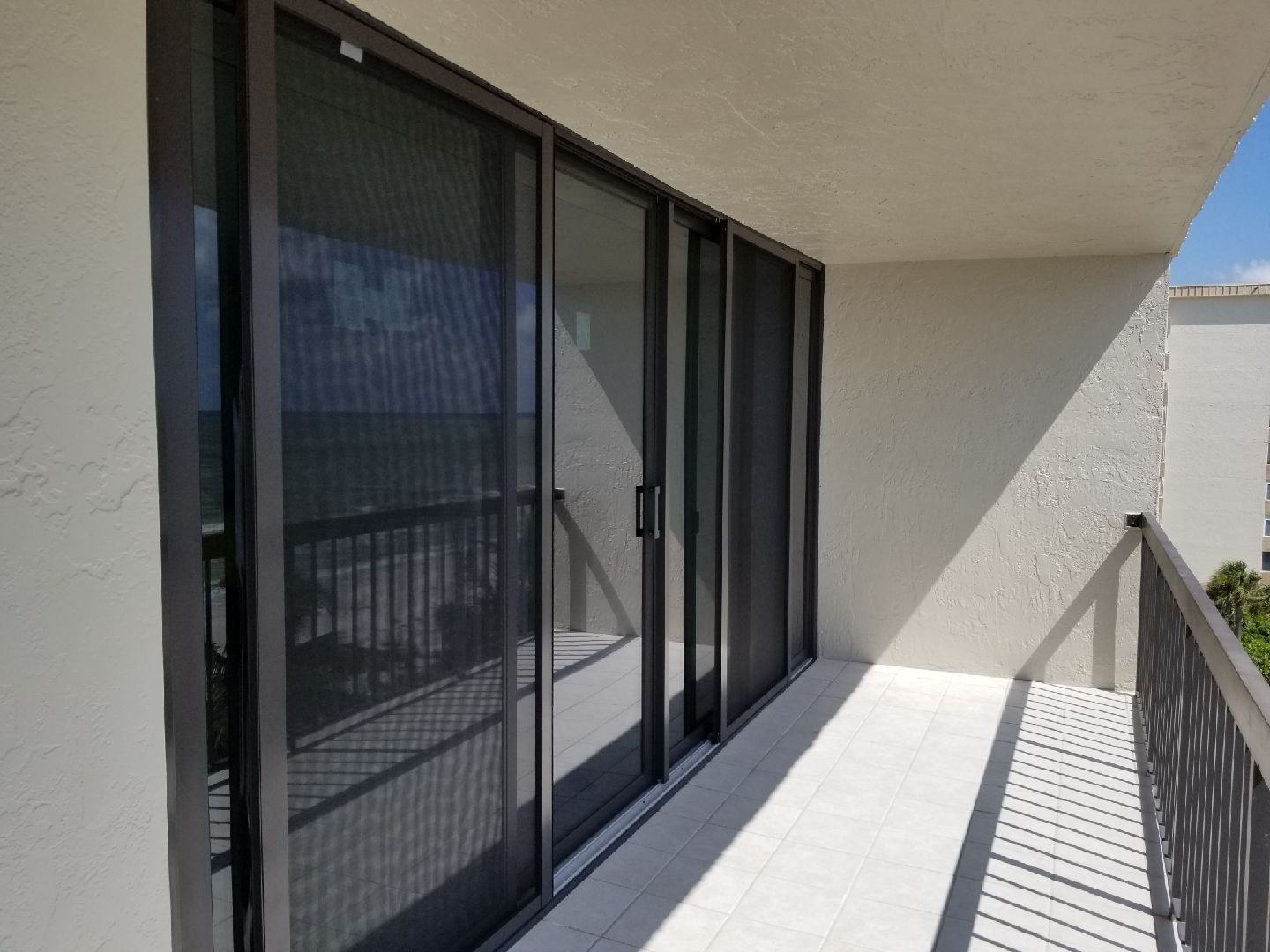 A sunny outdoor balcony featuring dark-framed sliding glass doors with screens, overlooking a coastal view.