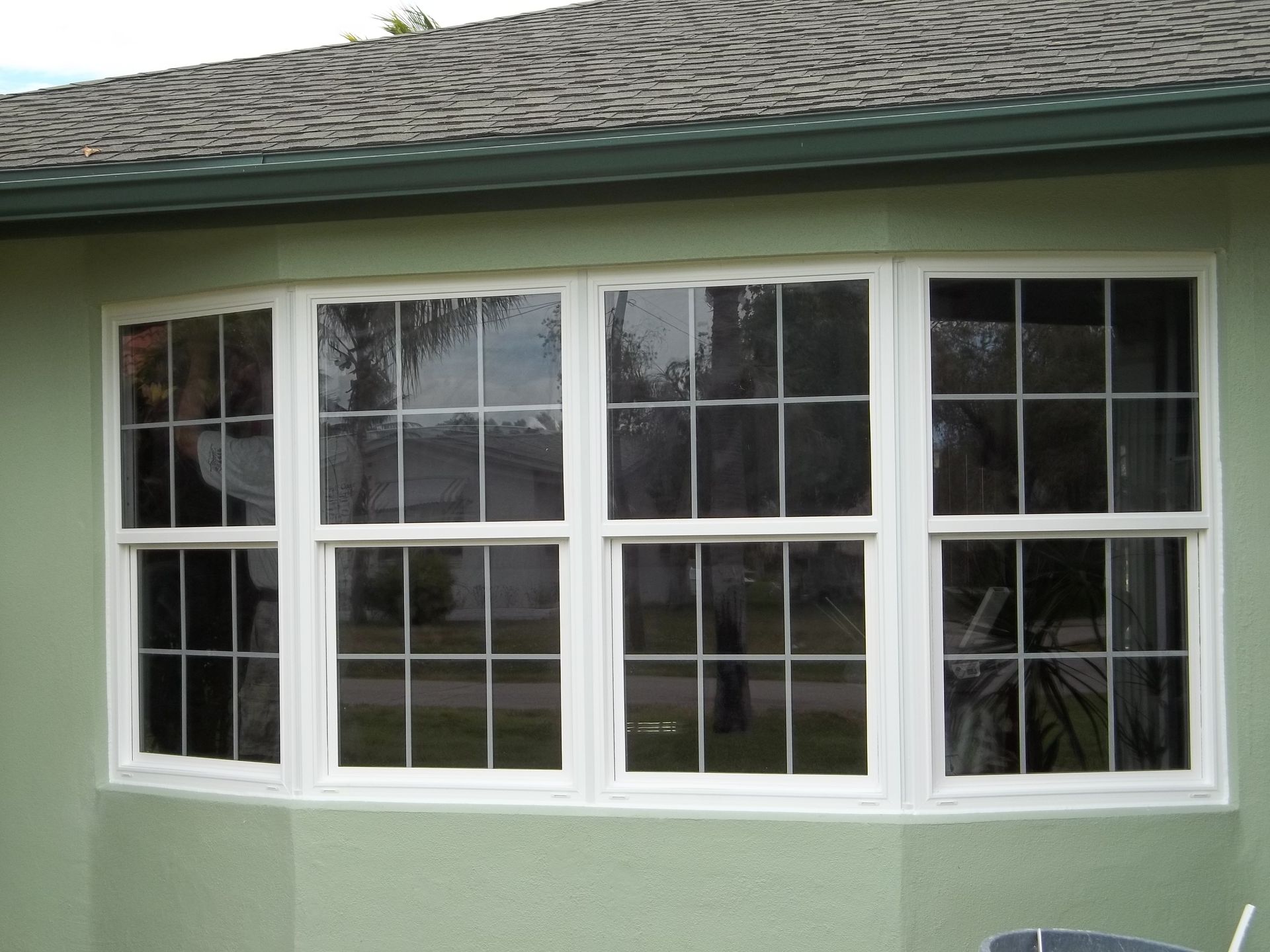 A four-section white bay window with a grid pattern, set into a light green exterior wall under a dark shingled roof.