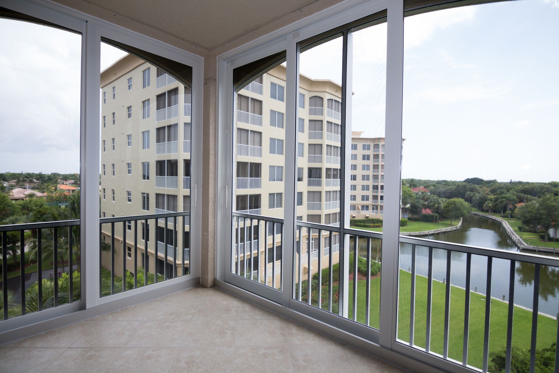 A bright indoor balcony with sliding glass doors looking out onto a residential building, green lawn, and a canal.