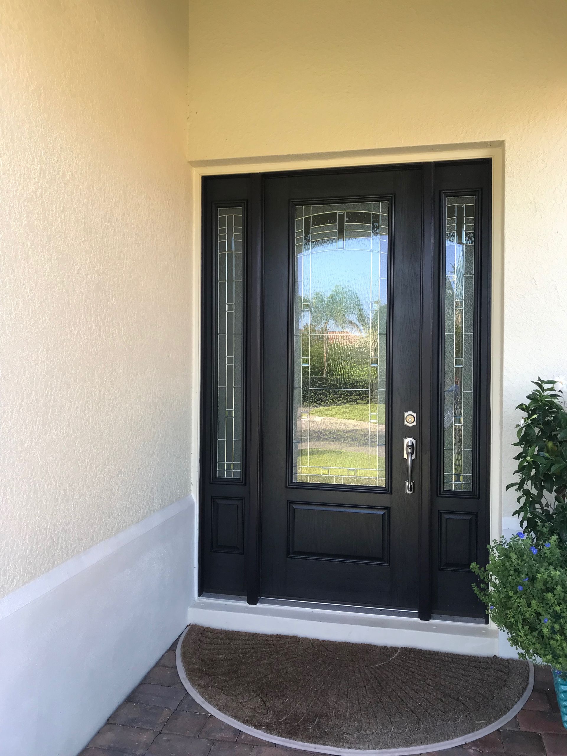 A black front door with a glass panel and two sidelights sits under a covered entryway with beige stucco walls.