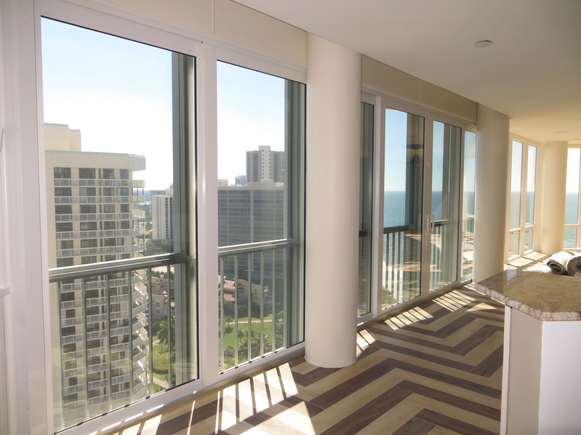 A high-rise room with floor-to-ceiling windows, chevron-patterned floors, and views of city buildings and the ocean.