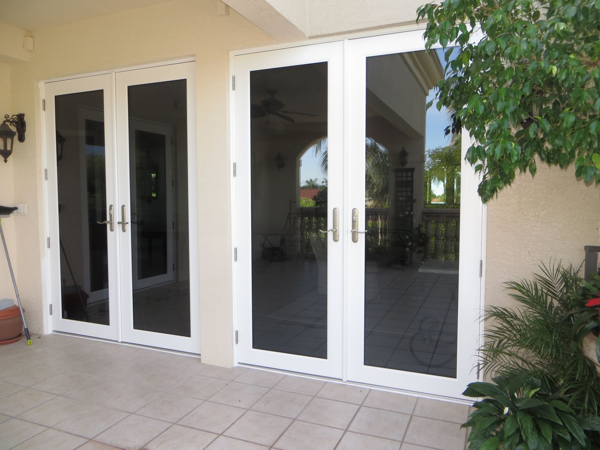 Two sets of white double glass doors with dark tint, installed on a tiled patio area next to a wall and a green plant.